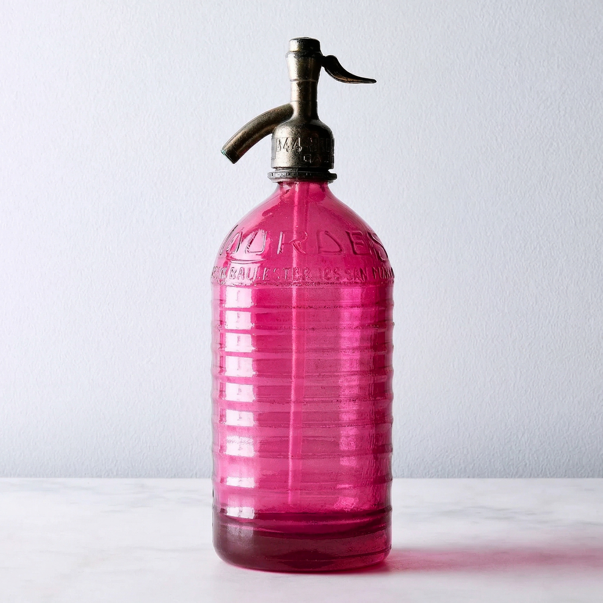 Vintage soda siphon in deep pink glass with a dark metal pump dispenser, standing on a light marble surface against a white wall.