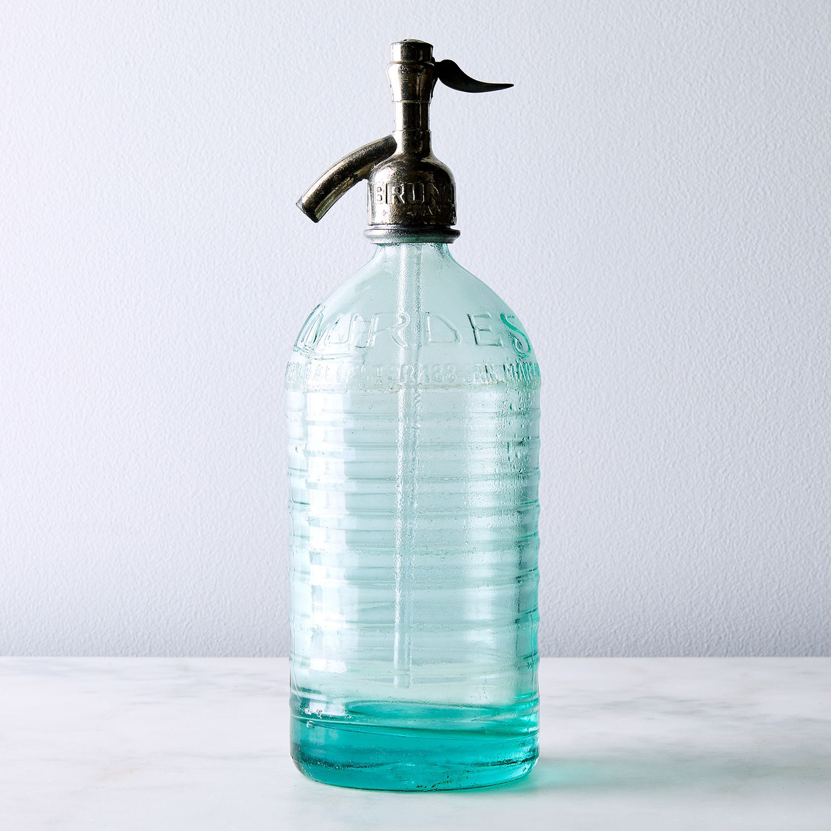  Clear Lourdes vintage Argentine seltzer bottle with ridged silhouette and embossed name. A soda bottle with a dark metal pump dispenser,  set against a light gray wall and white surface.